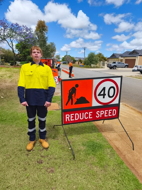 A teenager in hi-vis workwear beside a traffic-control sign on a Perth street